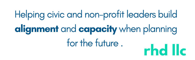 Three women sitting at a table in a conference room setting. One woman in a turquoise sweater is standing behind them, holding a paper and looking on with a smile.