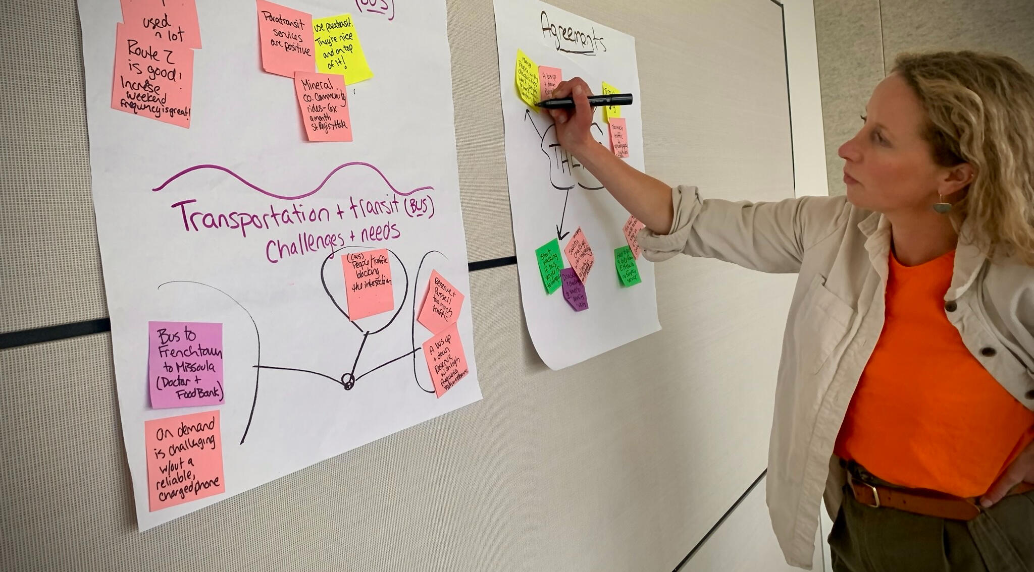 A white woman with blonde hair wearing a black shirt and brightly colored patterned scarf looks at the camera.