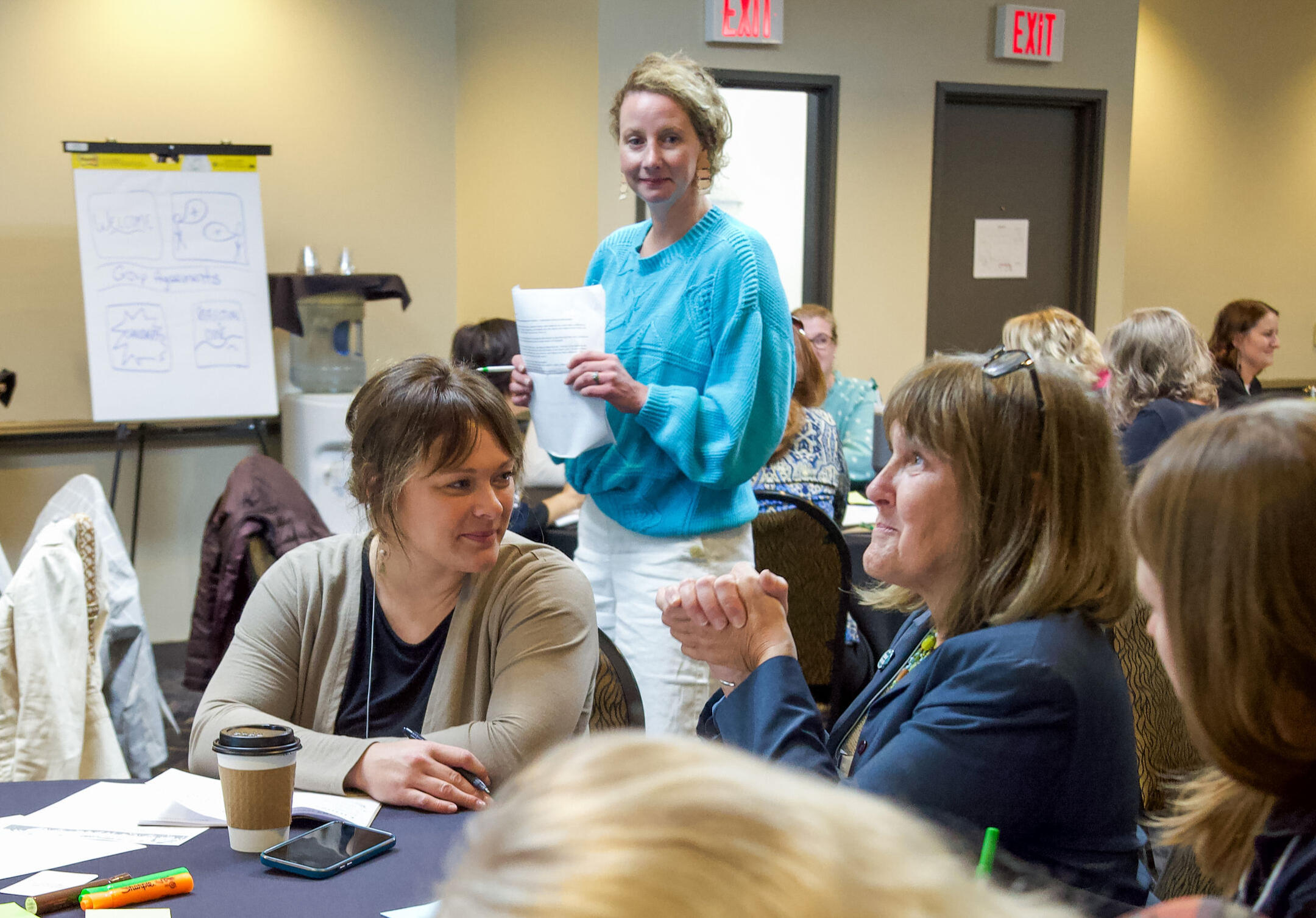 An adult women wearing a turquoise sweater stands and listens in on a round table of individuals in discussion, with two women in focus at a conference.