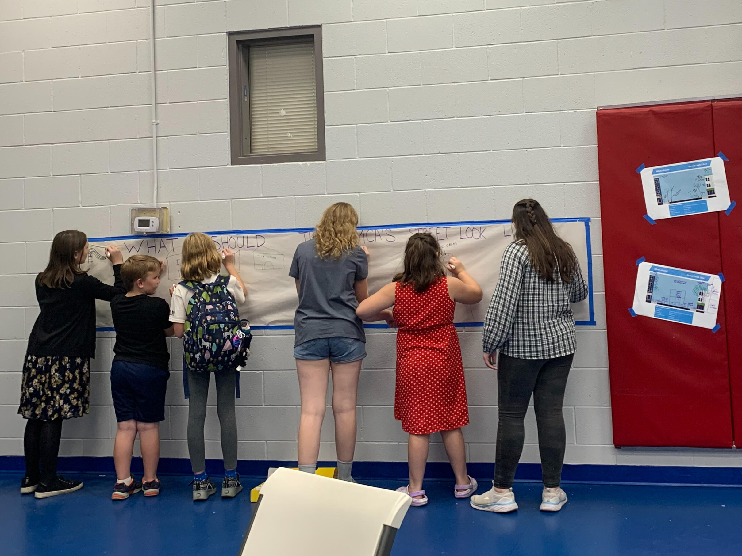 Six youth stand at a wall drawing ideas on a long sheet of paper.